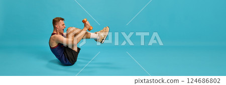 Athletic man holding dumbbells in boat pose, dressed in sportwear, working on core strength and balance, against blue background. 124686802