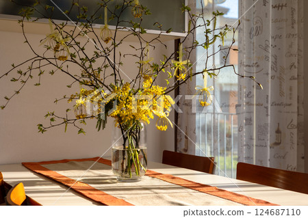 Festive table with bouquet of yellow mimosa and branches adorned with handmade eggs wire ornaments tied with yellow ribbon bows in glass vase. Decoration for Easter, Women's Day, or spring celebration Festive table with bouquet of yellow mimosa and branches adorned with handmade eggs wire ornaments tied with yellow ribbon bows in glass vase. Decoration for Easter, Women's Day, or spring celebration 124687110