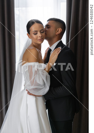 Groom kissing bride on cheek while embracing her near window 124688042