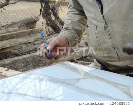 A worker's hand with a blue marker leans over a large glossy tile material on a workbench outside a house, marking trim tiles for cutting to the right size 124688935