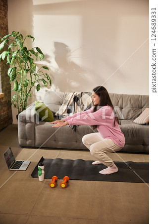 Girl in pink hoodie squatting on yoga mat in front of laptop, following workout instructions during home fitness session in cozy living room. 124689331
