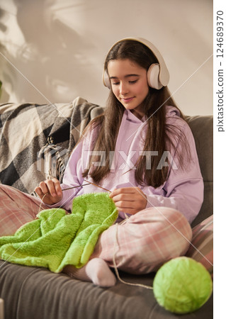 Girl focused on knitting green fabric in headphones while sitting on couch, engaged in creative home learning activity Girl focused on knitting green fabric in headphones while sitting on couch, engaged in creative home learning activity 124689370