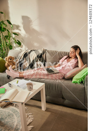 Teenage girl, student lying on couch with laptop, touching head in tiredness and boredom, attending online class, surrounded by notebooks and tea. Teenage girl, student lying on couch with laptop, touching head in tiredness and boredom, attending online class, surrounded by notebooks and tea. 124689372