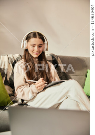 Smiling teen girl sitting on couch in headphones, attending online lesson at home, making notes on digital tablet with stylus. 124689404