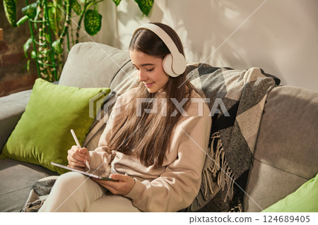 Teenage girl in headphones making notes on digital tablet while sitting on couch, fully immersed in creative home learning process. Teenage girl in headphones making notes on digital tablet while sitting on couch, fully immersed in creative home learning process. 124689405