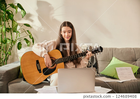 Beautiful. smiling teen girl sitting on couch at modern, stylish living room, playing guitar while looking at laptop screen during virtual music lesson Beautiful. smiling teen girl sitting on couch at modern, stylish living room, playing guitar while looking at laptop screen during virtual music lesson 124689408