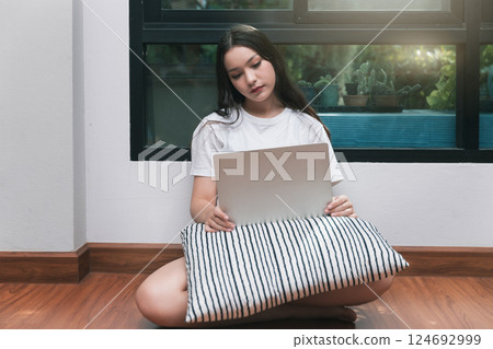 A young woman with laptop and pillow sitting drowsy on the living room floor. 124692999