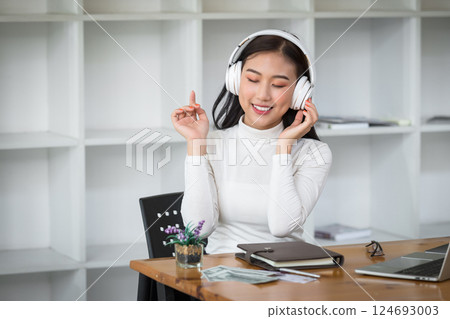 Asian woman wearing headphones listening to music on her laptop. Relaxing time. Lazy worker wasting time at workplace in co-working. 124693003
