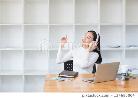 Asian woman wearing headphones listening to music on her laptop. Relaxing time. Lazy worker wasting time at workplace in co-working. 124693004