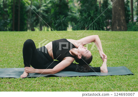 Asian woman practicing yoga in Revolved Head-to-Knee Pose on the mat in outdoor park. 124693007