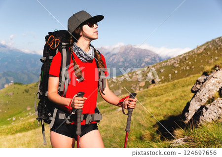 Portrait of a tourist. Female tourist happy and smiling during a hike. Beautiful young Caucasian sports model joyful, standing and looking at the route with a backpack and trekking poles Portrait of a tourist. Female tourist happy and smiling during a hike. Beautiful young Caucasian sports model joyful, standing and looking at the route with a backpack and trekking poles 124697656