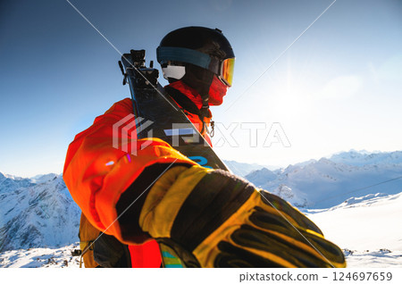 guy in a ski suit holds skis in his hand and stands on a snowy mountain looking into the distance at the sun and a mountain range, ski resort and recreation development 124697659
