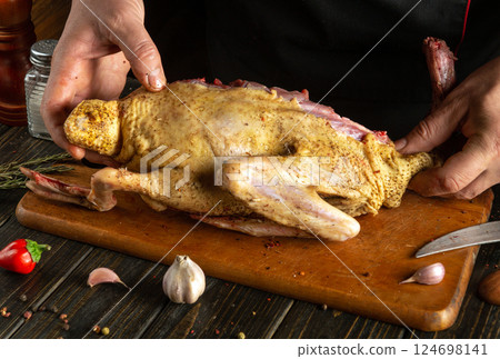 A man holds a whole duck seasoned with spices while cooking it on a wooden cutting board in a cozy kitchen. The decor includes fresh garlic, herbs and chili peppers 124698141