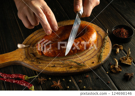 A person slices a flavorful sausage placed on a rustic wooden cutting board. The setting features various herbs and spices, enhancing the culinary experience A person slices a flavorful sausage placed on a rustic wooden cutting board. The setting features various herbs and spices, enhancing the culinary experience 124698146