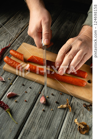 Hands expertly slice through savory sausages and garlic on a wooden cutting board. The kitchen has a warm atmosphere, enhanced by nearby spices and herbs Hands expertly slice through savory sausages and garlic on a wooden cutting board. The kitchen has a warm atmosphere, enhanced by nearby spices and herbs 124698158
