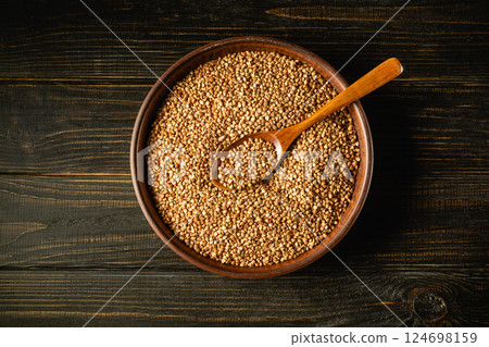 A wooden bowl filled with buckwheat grains sits on a dark wooden table. A wooden spoon sits among the buckwheat seeds, showing their texture and color in natural light. A wooden bowl filled with buckwheat grains sits on a dark wooden table. A wooden spoon sits among the buckwheat seeds, showing their texture and color in natural light. 124698159