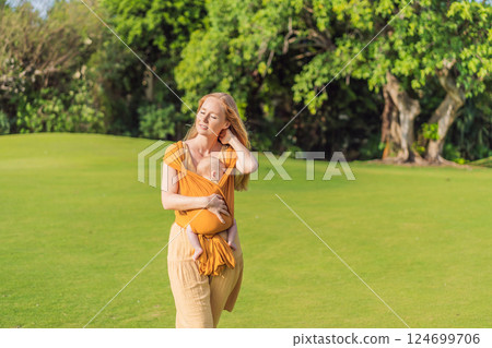 Mother holding her little baby in a yellow sling in the park. Warm and loving family moment. Babywearing, parenting, and mother-child bonding concept Mother holding her little baby in a yellow sling in the park. Warm and loving family moment. Babywearing, parenting, and mother-child bonding concept 124699706