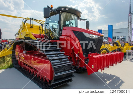 A large red tracked agricultural vehicle with a cabin and various attachments on display at an outdoor machinery exhibition 124700014