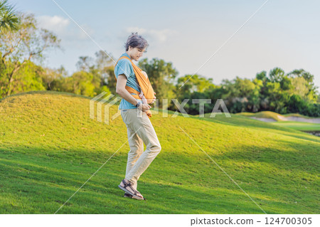 Father holding his little baby in a yellow sling in the park. Warm and loving family moment. Babywearing, fatherhood, and parent-child bonding concept 124700305