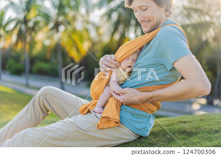 Father holding his little baby in a yellow sling in the park. Warm and loving family moment. Babywearing, fatherhood, and parent-child bonding concept 124700306