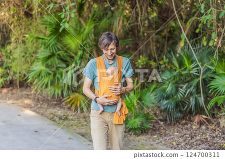 Father holding his little baby in a yellow sling in the park. Warm and loving family moment. Babywearing, fatherhood, and parent-child bonding concept Father holding his little baby in a yellow sling in the park. Warm and loving family moment. Babywearing, fatherhood, and parent-child bonding concept 124700311
