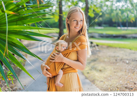 Mother holding her little baby in a yellow sling in the park. Warm and loving family moment. Babywearing, parenting, and mother-child bonding concept Mother holding her little baby in a yellow sling in the park. Warm and loving family moment. Babywearing, parenting, and mother-child bonding concept 124700313