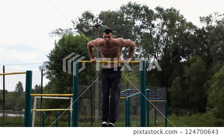 Hardy shirtless sportsman doing push ups on horizontal bar at nature. Strong and muscular guy training at sports ground. Young athletic guy exercising outdoor. Concept of active and healthy lifestyle 124700643