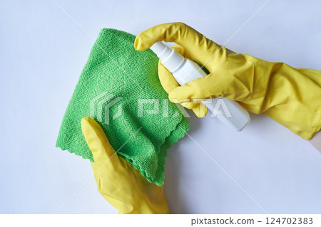Person wearing yellow gloves cleaning with green cloth and spray bottle on white 124702383