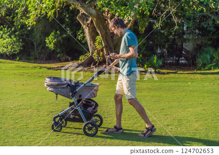 Father pushing a baby stroller on a grassy field while looking at his phone. Concept of modern parenting, multitasking, technology in daily life, and work-life balance 124702653