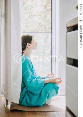Young woman meditating in lotus position by a bright window with eyes closed. New Age wellness for Gen Z and Millennials, spiritual reset, conscious calm, incense culture 124708885