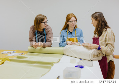 Three female seamstresses fashion designers stand at a table with roll of fabric and negotiate. Three female seamstresses fashion designers stand at a table with roll of fabric and negotiate. 124715234