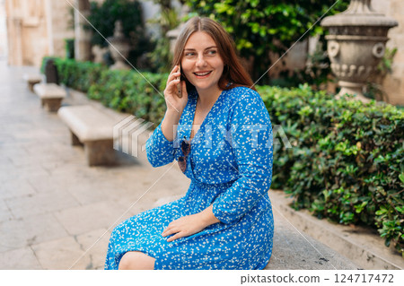 Beautiful stylish woman in trendy wear spending time on street using smartphone. Happy girl in dress talking on the mobile phone. Urban lifestyle concept, old town 124717472