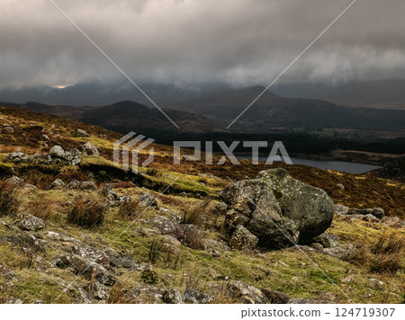 Landscape featuring a field with rocks, rolling hills, and a cloudy sky above. Landscape featuring a field with rocks, rolling hills, and a cloudy sky above. 124719307