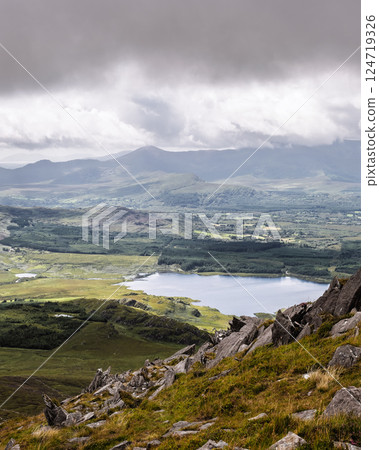 High-angle view of a serene landscape. A lake is visible nestled within rolling hills and valleys, surrounded by varied vegetation. Grey rocks are in foreground. 124719326
