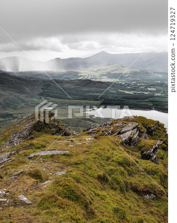 Panoramic view from an elevated position. The image shows a lake in a green valley, surrounded by hills and distant mountains under a light overcast sky. 124719327