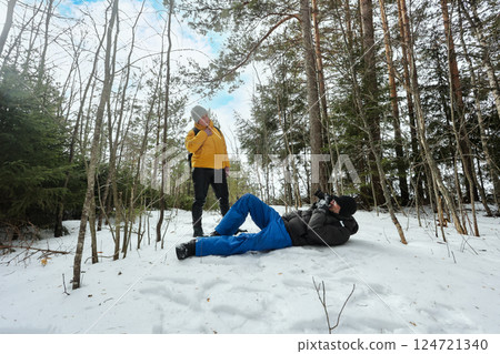Mature couple pauses during hike as man takes photo lying on ground and woman stands in front of him enjoying playful moment in quiet snowy forest. 124721340