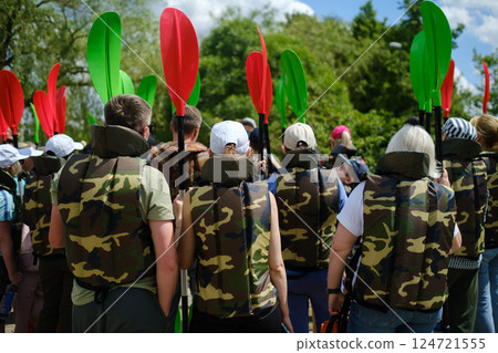 Group of individuals preparing for kayaking activity at a lake surrounded by greenery on a sunny day with colorful paddles visible Group of individuals preparing for kayaking activity at a lake surrounded by greenery on a sunny day with colorful paddles visible 124721555