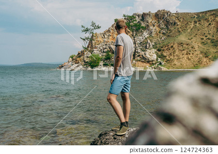 man stands on stone with his back to camera, on shore of lake with rocky cliffs man stands on stone with his back to camera, on shore of lake with rocky cliffs 124724363