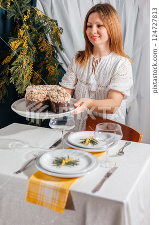 Woman in a white dress sets a festive table with Traditional Easter Colomba and mimosa. Spring mood and cozy atmosphere. 124727813