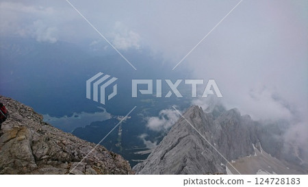 Zugspitze, Germany - 6.17.2018: Breathtaking view from the summit overlooking a dramatic landscape with rugged peaks shrouded in mist and Eibsee reflecting the cloudy sky Zugspitze, Germany - 6.17.2018: Breathtaking view from the summit overlooking a dramatic landscape with rugged peaks shrouded in mist and Eibsee reflecting the cloudy sky 124728183
