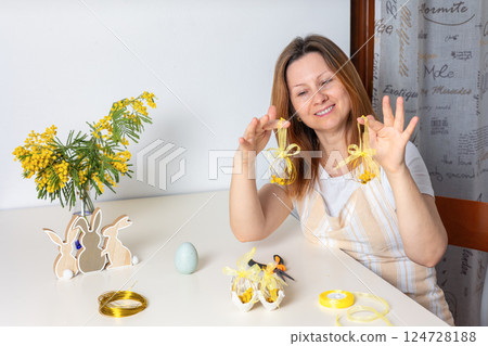 Smiling woman making handmade Easter egg wire ornaments with mimosa and yellow ribbon at a festive spring table. Creative holiday crafts and DIY Easter decoration. 124728188