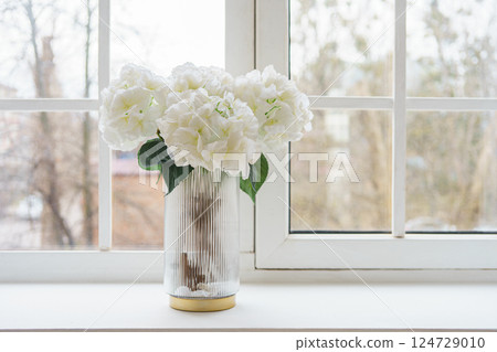 Bouquet of white artificial decorative hydrangea, hortensia in vase on windowsill in living room 124729010