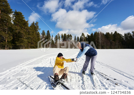 Mature couple enjoys skiing on sunny winter day, middle aged man in yellow struggling to get up from snow while woman offers assistance, forest background. Mature couple enjoys skiing on sunny winter day, middle aged man in yellow struggling to get up from snow while woman offers assistance, forest background. 124734836
