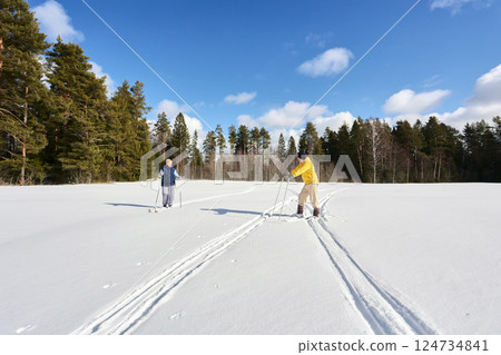 Individuals pause during cross country skiing, one in yellow adjusts equipment, while other stands with poles, both on snow covered ground under open sky. Individuals pause during cross country skiing, one in yellow adjusts equipment, while other stands with poles, both on snow covered ground under open sky. 124734841