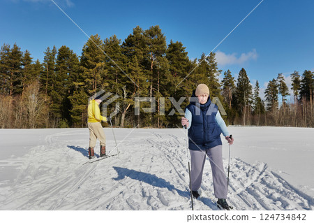 Middle aged couple enjoys cross country skiing, woman skiing towards camera while man trails, both gliding over snow with poles in sunny open field. Middle aged couple enjoys cross country skiing, woman skiing towards camera while man trails, both gliding over snow with poles in sunny open field. 124734842