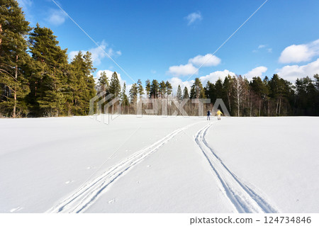 Distant figures ski across snow covered field, one wearing bright yellow clothing while other follows, both moving toward distant treeline on sunny day. Distant figures ski across snow covered field, one wearing bright yellow clothing while other follows, both moving toward distant treeline on sunny day. 124734846