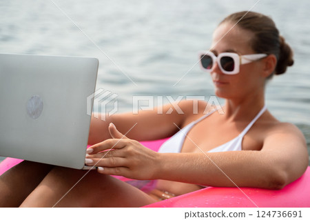 A woman is sitting in a pink inflatable raft on a lake, holding a laptop. Concept of relaxation and leisure, as the woman enjoys her time in the water while working on her laptop. 124736691