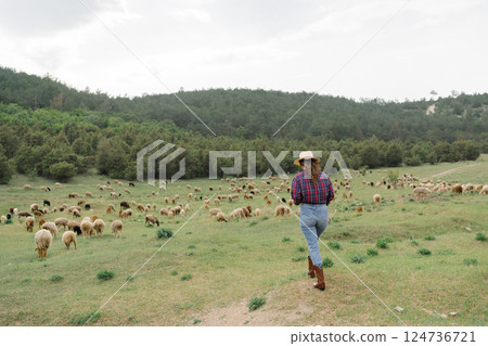 A woman wearing a cowboy hat is walking through a field of sheep 124736721