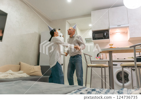Couple Dancing Birthday Apartment: Happy senior couple joyfully dances wearing party hats in their modern apartment during birthday celebration. 124736735