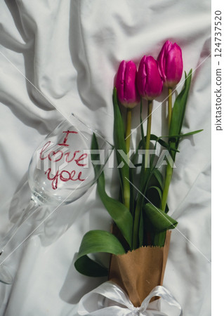 Bouquet of pink tulips on messy bed. I LOVE YOU written text on wine glass. Romantic morning scene. Greeting card Holiday flowers springtime 124737520
